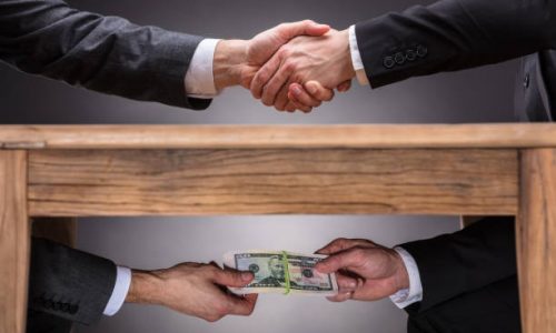 Close-up Of Two Businesspeople Shaking Hand And Taking Bribe Under Wooden Table On Grey Background